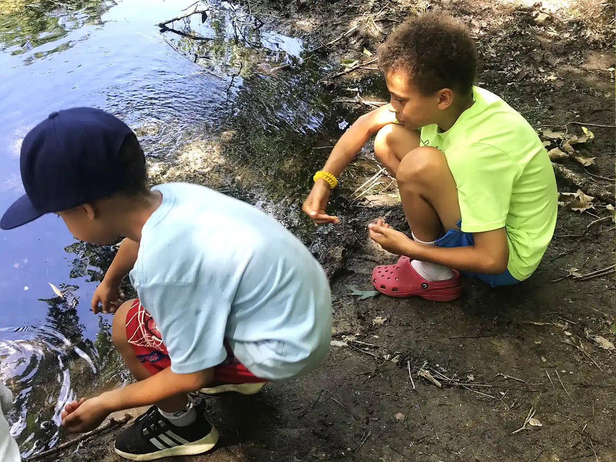 Two boys crouching by a body of water outdoors. The boy on the left wears a navy blue baseball cap, a light blue t-shirt, and red plaid shorts, while the boy on the right wears a bright lime green t-shirt, blue shorts, pink Crocs, and a yellow wristband.