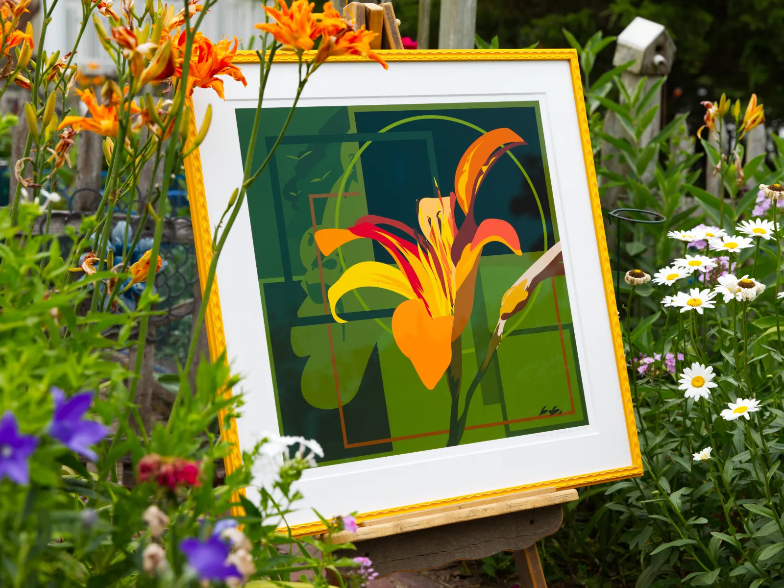 A framed floral art print featuring a stylized orange and yellow lily on a dark green geometric background, displayed on an easel in a garden surrounded by real flowers and foliage.