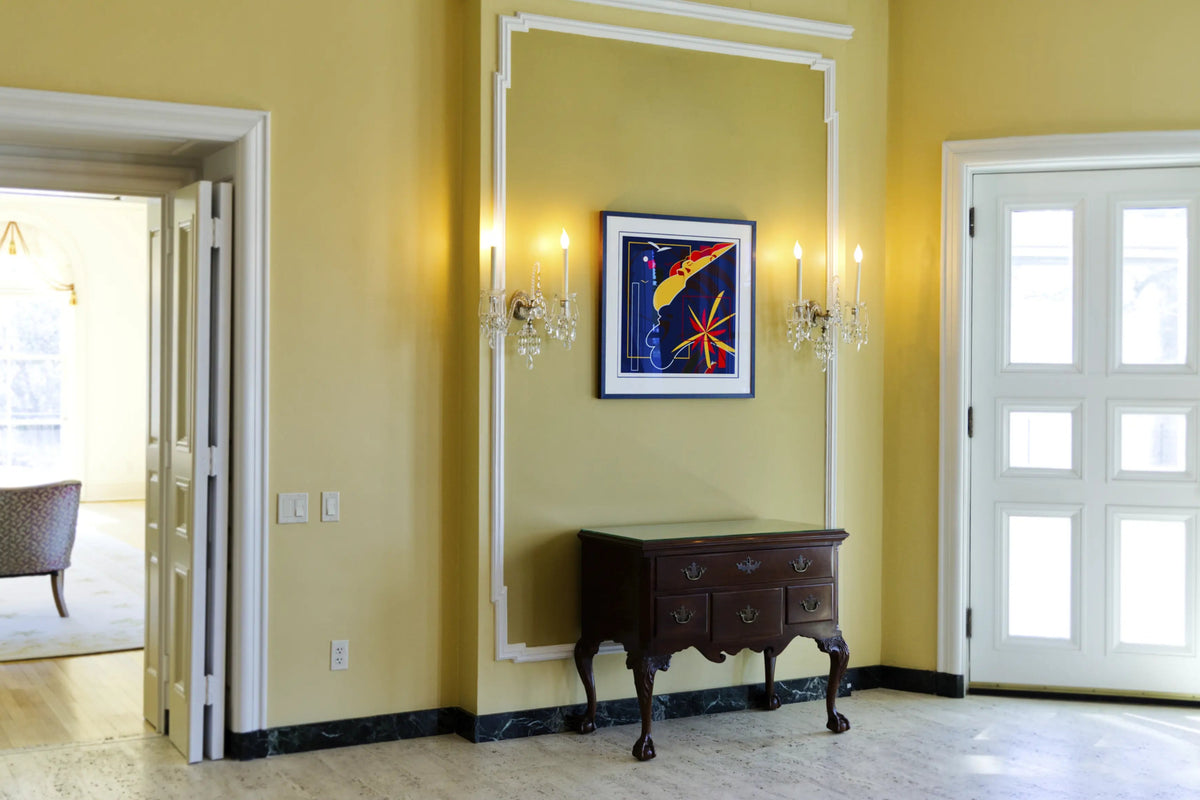 A framed abstract art print with a blue and red starburst design hangs on a mustard yellow wall in a hallway, flanked by two lit crystal wall sconces above a dark wood console table. White molding frames the wall art area, with an open doorway on the left and a white door on the right.