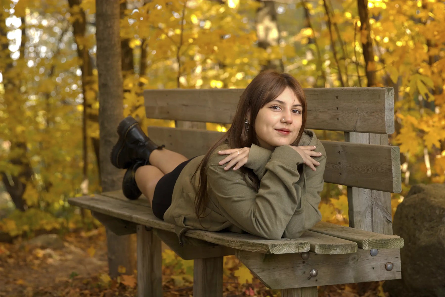 A woman with long brown hair lies on a wooden park bench in an autumn forest, wearing an olive green jacket, a black skirt, and black boots. The background is filled with golden yellow leaves.