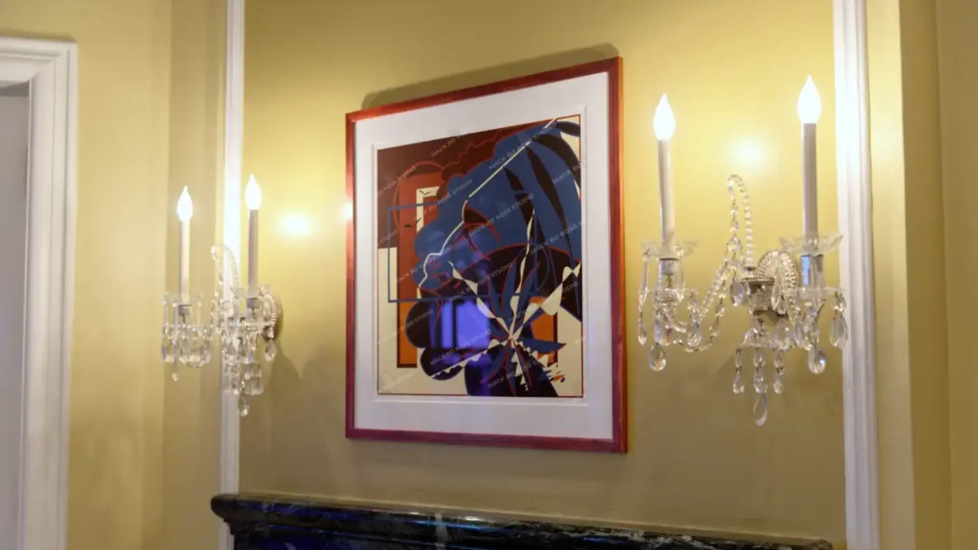 Framed abstract art on a yellow wall with crystal sconces.