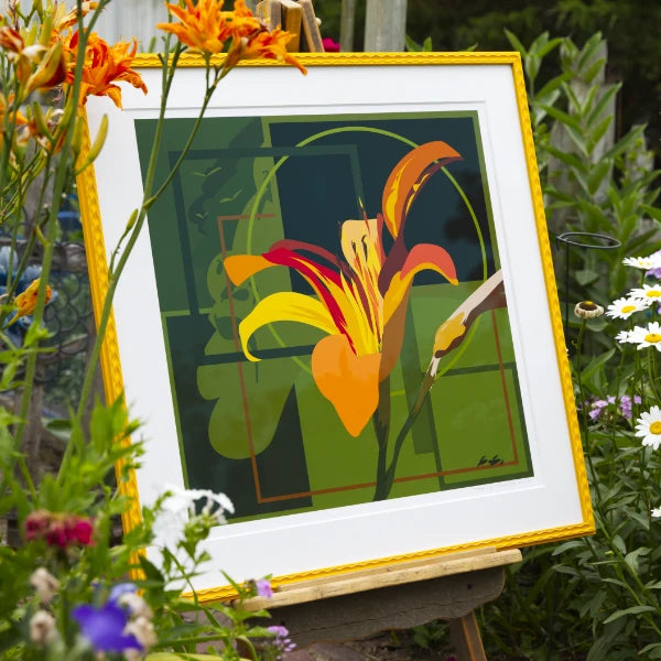 A framed art print featuring a stylized orange and yellow lily flower on a green geometric background, displayed on an easel in a garden setting with flowers around it.
